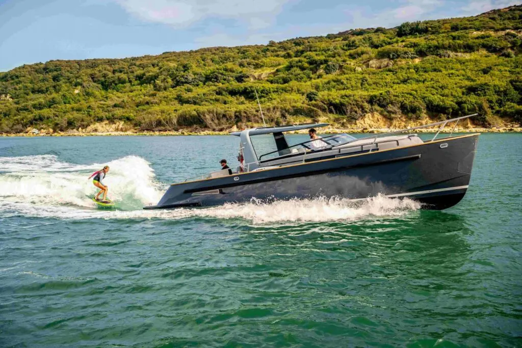 Duchy Sport with a dark hull towing a wakesurfer along a wooded estuary shoreline