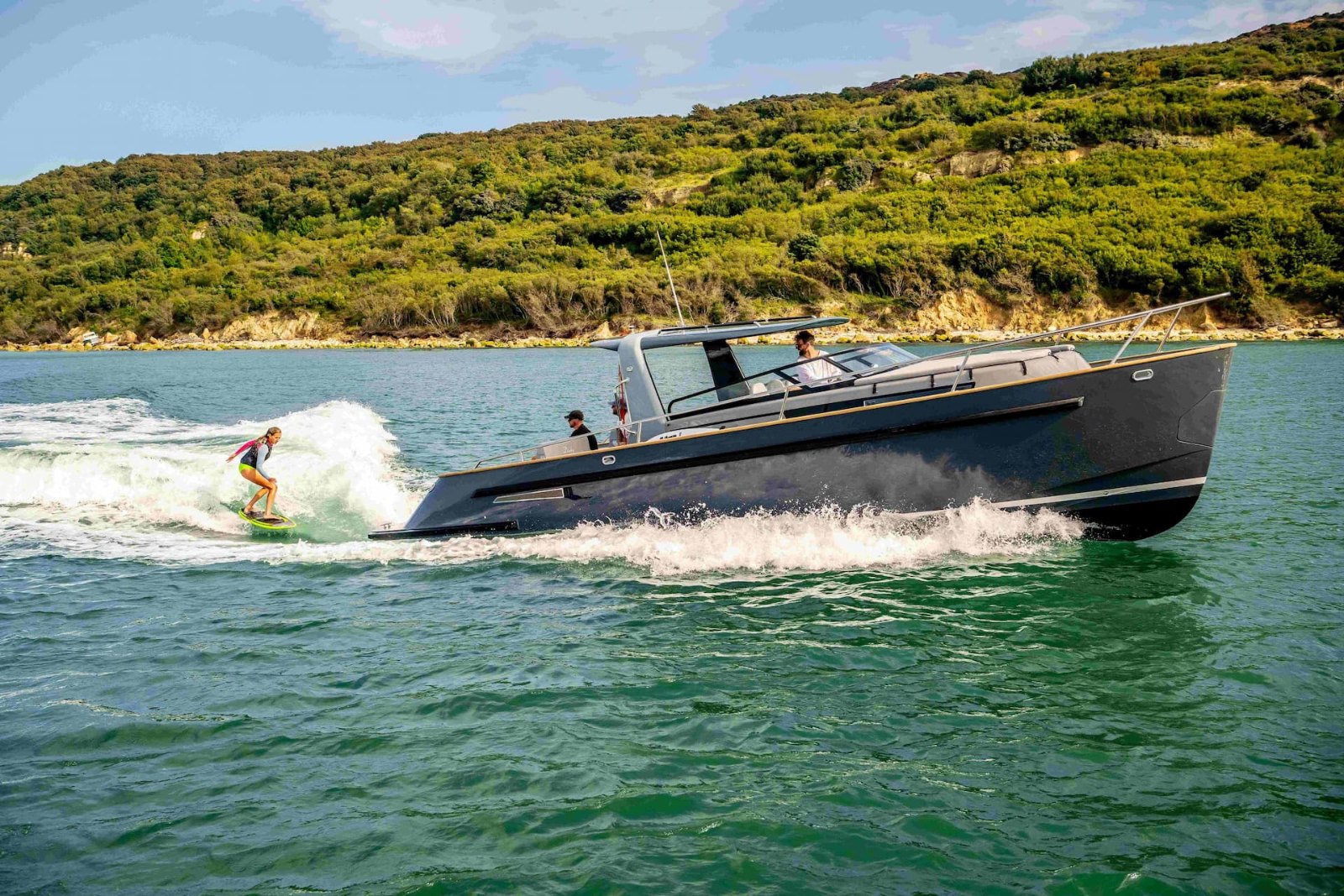 Duchy Sport with a dark hull towing a wakesurfer along a wooded estuary shoreline
